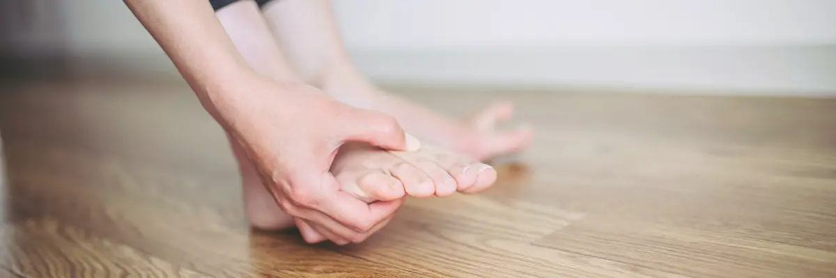 フローリングの床の上で、女性が自分の足の指の付け根（つま先付近）を両手で押さえ、痛みを確認している様子の写真。モートン病の症状や原因を解説するページのトップ画像。