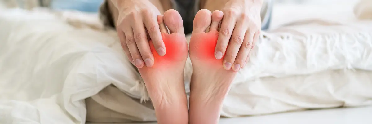 ベッドの上で両足の裏を見せ、中足骨頭（足の指の付け根の関節）のあたりが赤く痛みのイメージとして強調されている写真。中足骨頭痛の症状や原因を解説するメインビジュアル。